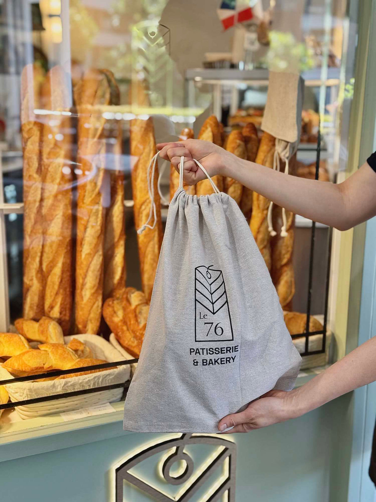 A Le 76 cloth bag held in front of the shop window, with baguettes and pastries on the shelves behind
