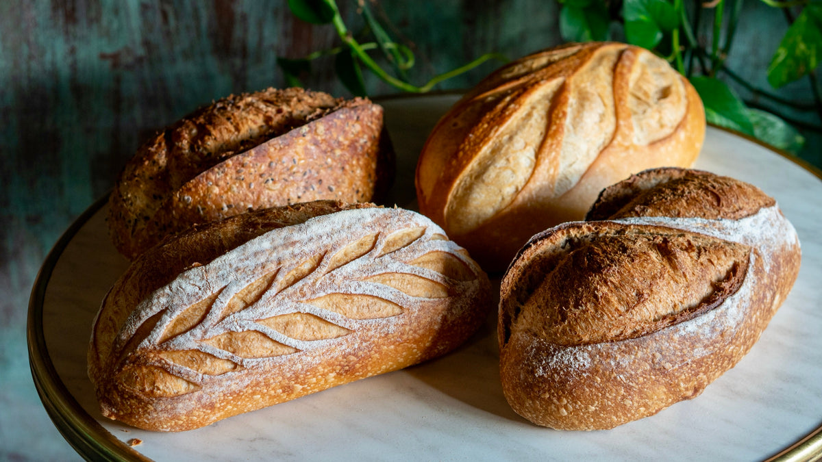 Four scored sourdough loaves on a marble platter, one dusted with flour in a wheat-ear pattern